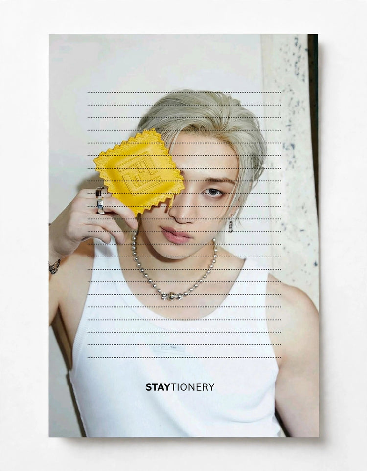 A vertical stationery card or notebook cover featuring a portrait of a person with platinum blonde hair, wearing a white tank top and silver jewelry, holding a yellow square card in front of one eye. The card has horizontal dashed lines and the word 'STAYTIONERY' printed at the bottom.