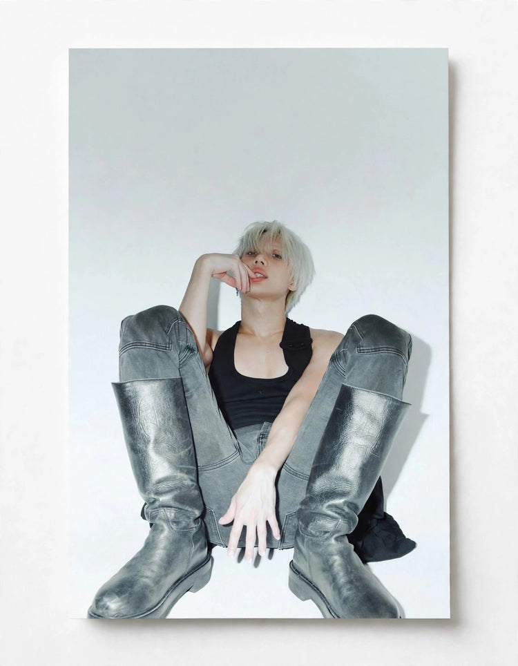 A vertical photographic print of a model with platinum blonde hair sitting cross-legged, wearing a black tank top, gray pants, and metallic silver knee-high boots, displayed on a light gray wall.