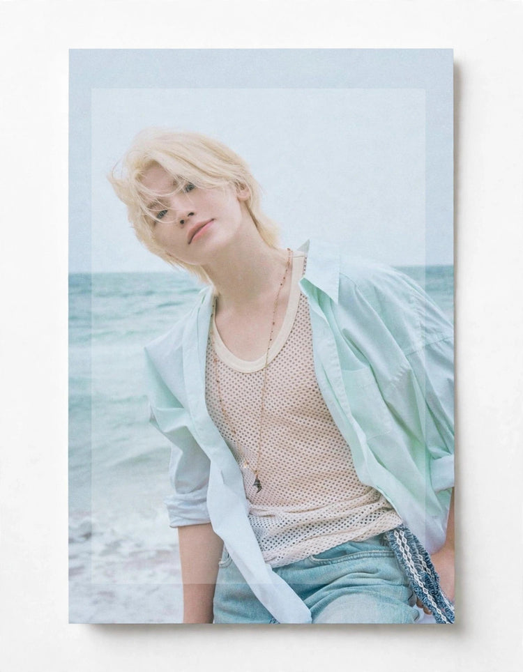 A vertical photographic print or poster showing a blonde person leaning against a beach background, wearing a pale mint shirt over a beige mesh top and light blue jeans. The print is photographed against a plain white wall.